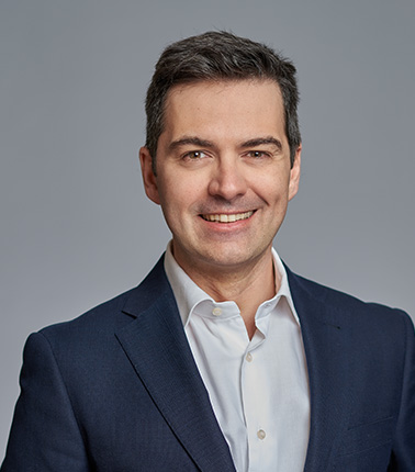 Uomo in giacca blu e camicia bianca su sfondo grigio chiaro.