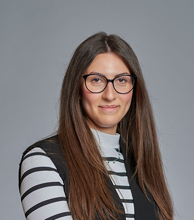 Donna con occhiali, capelli lunghi, vestita con maglione a righe e gilet scuro.