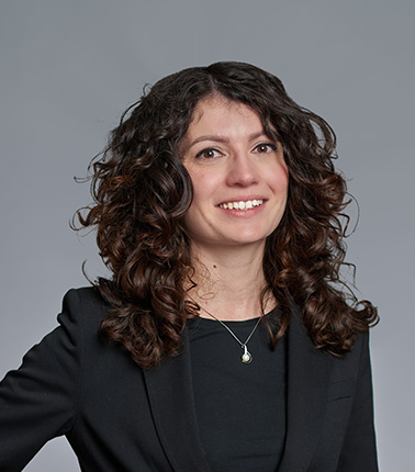 Donna con capelli ricci e abito nero su sfondo grigio.