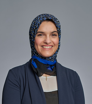 Donna sorridente con hijab blu e giacca scura su sfondo grigio.