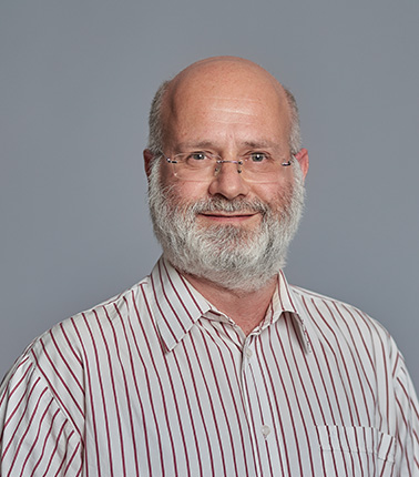 Uomo calvo con barba, occhiali e camicia a righe su sfondo grigio.