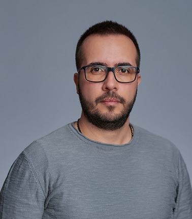 Uomo con occhiali e barba corta indossa una maglia grigia su sfondo grigio.