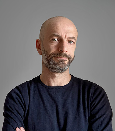 Uomo calvo con barba e maglietta scura su sfondo grigio.