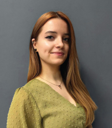 Giovane donna con capelli castani e maglia verde davanti a sfondo grigio.