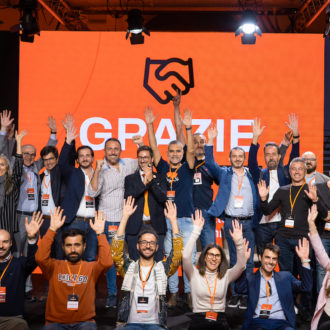 Gruppo di persone sorridenti su un palco con schermo arancione dietro che dice 'GRAZIE'.